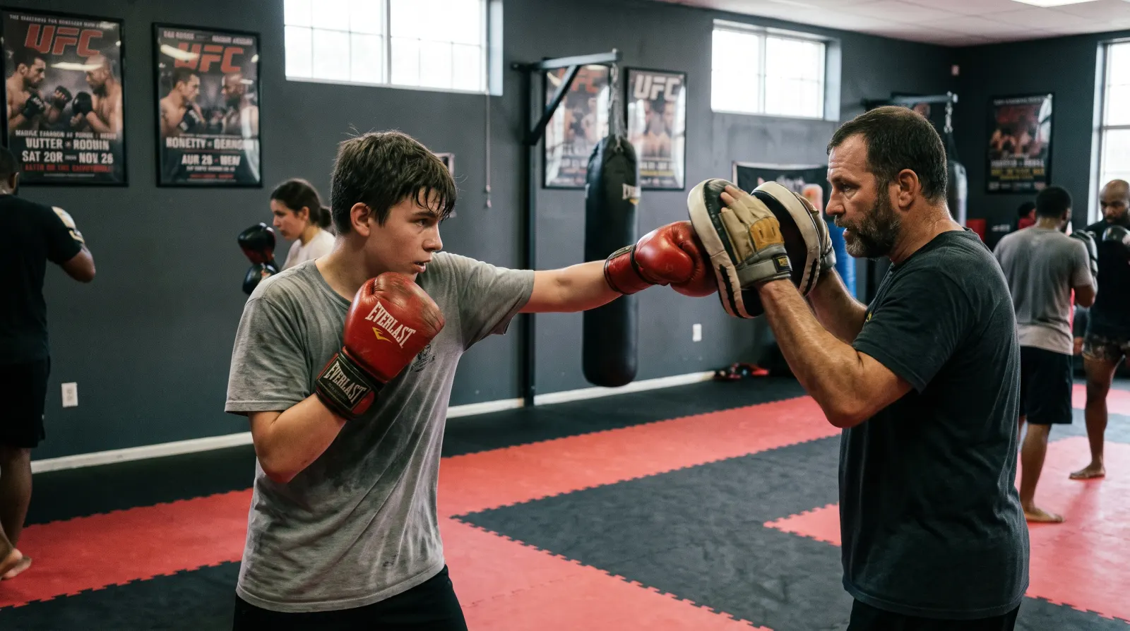 A teenage boy in boxing gloves throwing a jab at focus mitts held by a coach on red and black mats at a Northern Virginia boxing gym, with heavy bags and UFC posters in the background