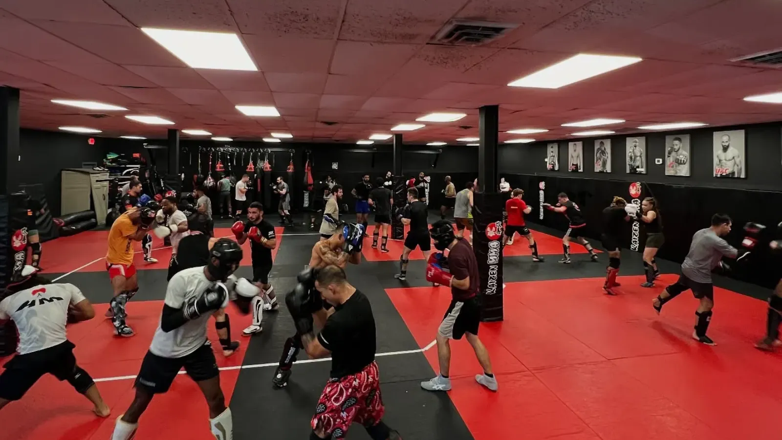 A full adult striking class at Kaizen MMA working in pairs across red and black mats, students in gloves and shin guards drilling combinations while heavy bags hang along the dark grey walls under UFC posters