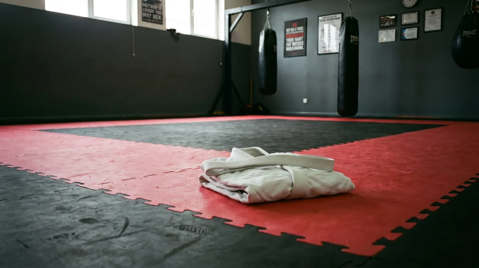 A folded white Brazilian jiu-jitsu gi with a white belt resting on red and black training mats inside a Northern Virginia jiu-jitsu academy, heavy bags and posters on dark grey walls in the background