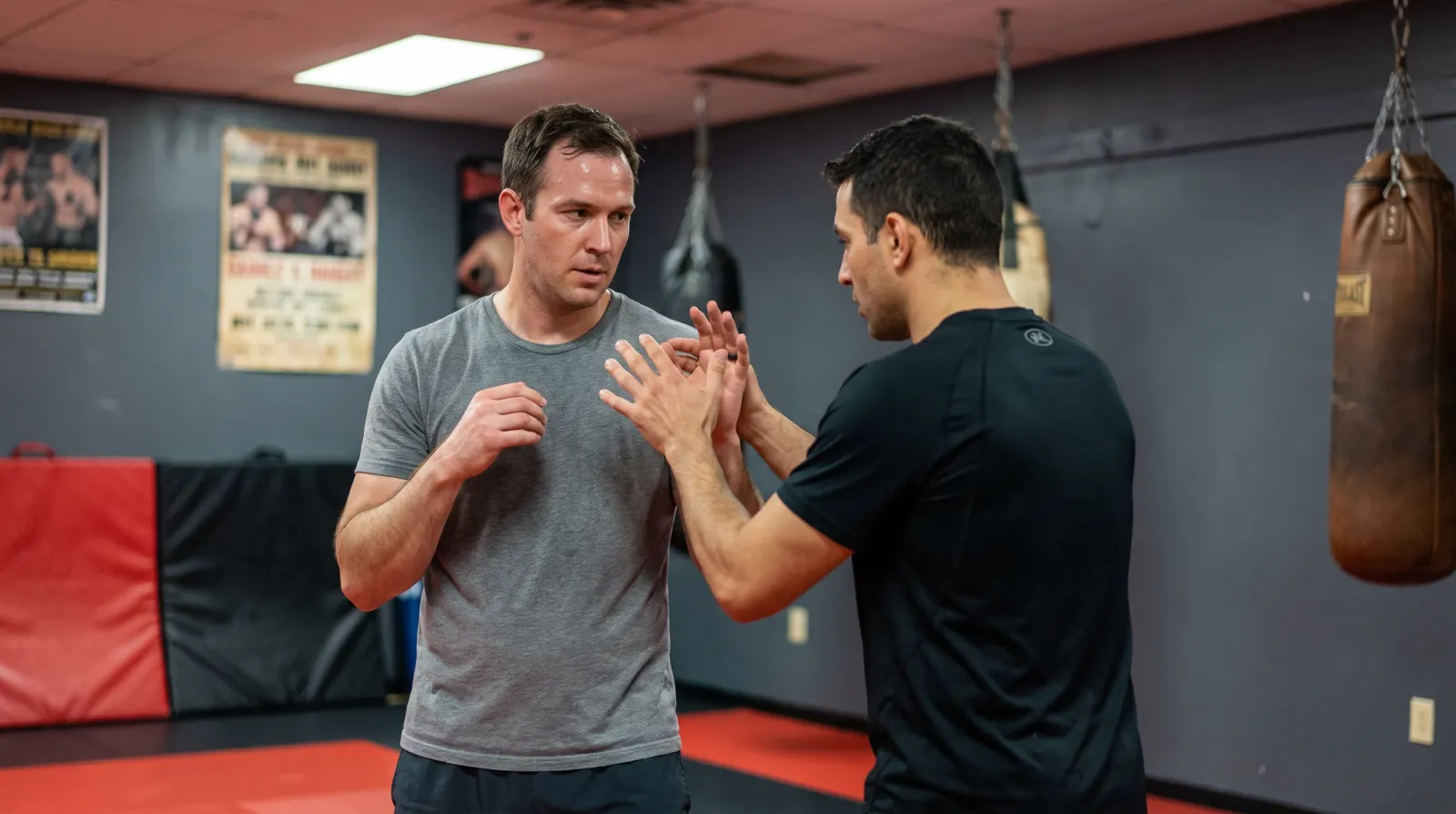 An average-build adult beginner in a gray t-shirt learning how to hold his hands up from a patient coach on red and black mats at a Northern Virginia martial arts gym, with heavy bags and UFC posters blurred in the background