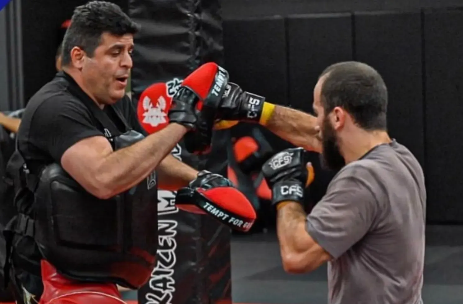 A Kaizen MMA coach in a body protector holding red focus mitts while a student in a grey t-shirt and gloves punches into the pad during a Northern Virginia striking class, a Kaizen-branded kick shield and dark grey gym walls behind them
