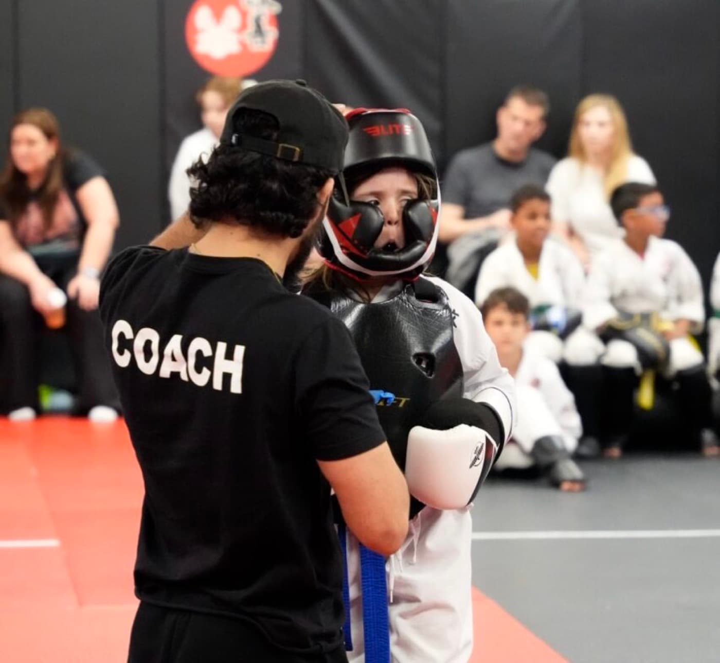A Kaizen MMA South Riding coach working one-on-one with a young Little Lion in safety gear while parents watch