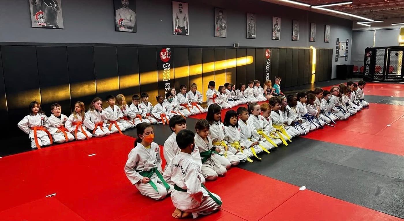 The Little Lions class of young kids ages 4 to 7 in white gis lined up on the mats at Kaizen MMA South Riding